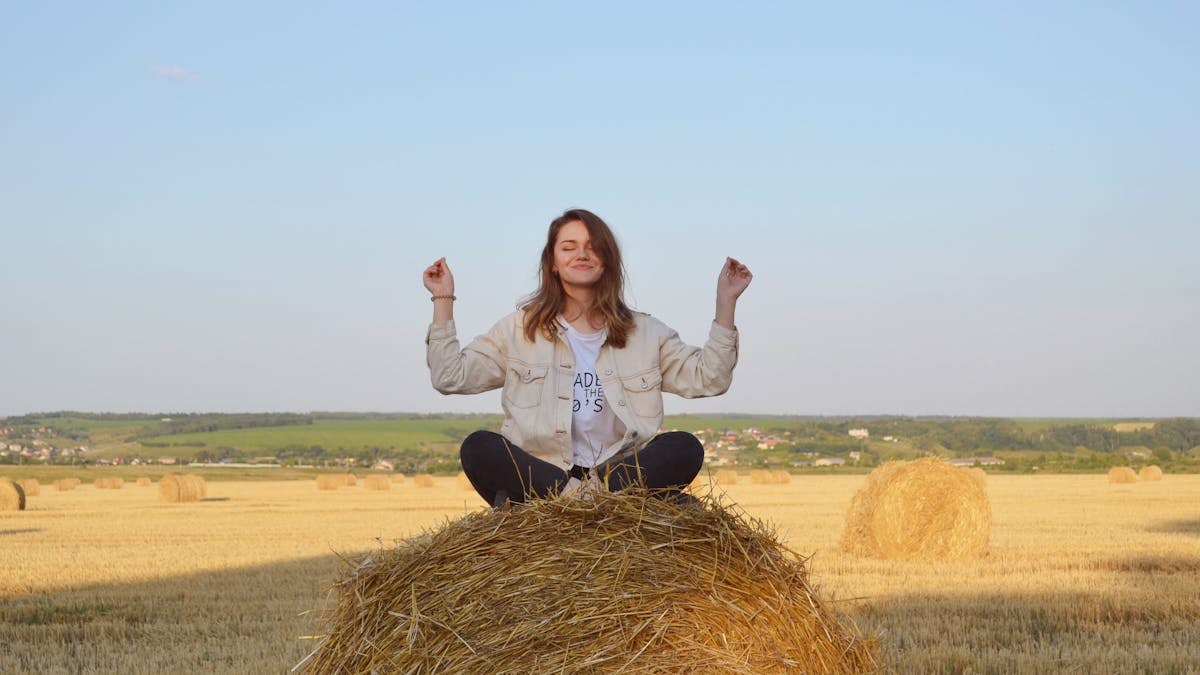 A young woman sits peacefully on a hay bale in a serene summer field.